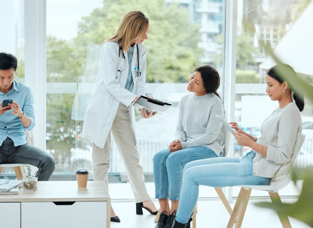 A person in a doctor's coat consults with another person while two people nearby are using electronic devices in a bright room.