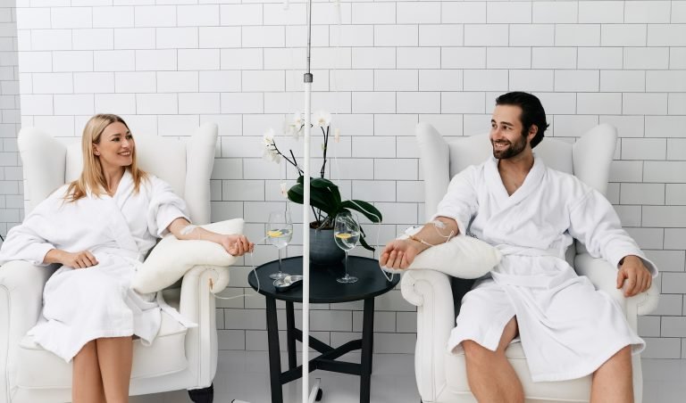 Two people in white robes relax in armchairs, receiving IV drips. A small table with drinks and a plant separates them.