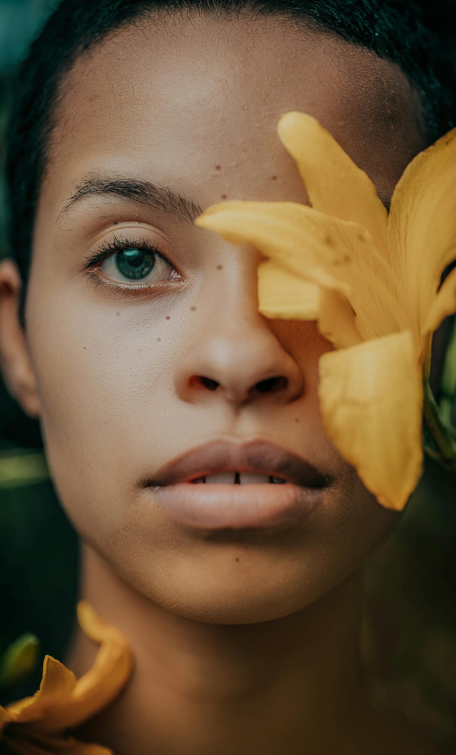 A person gazes forward with a yellow flower partially covering one eye, creating an artistic and serene close-up portrait.