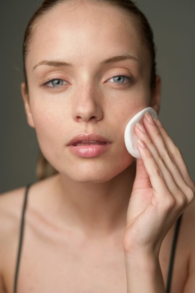 A person is cleansing their face with a cotton pad, focused expression, natural lighting, minimalist background.