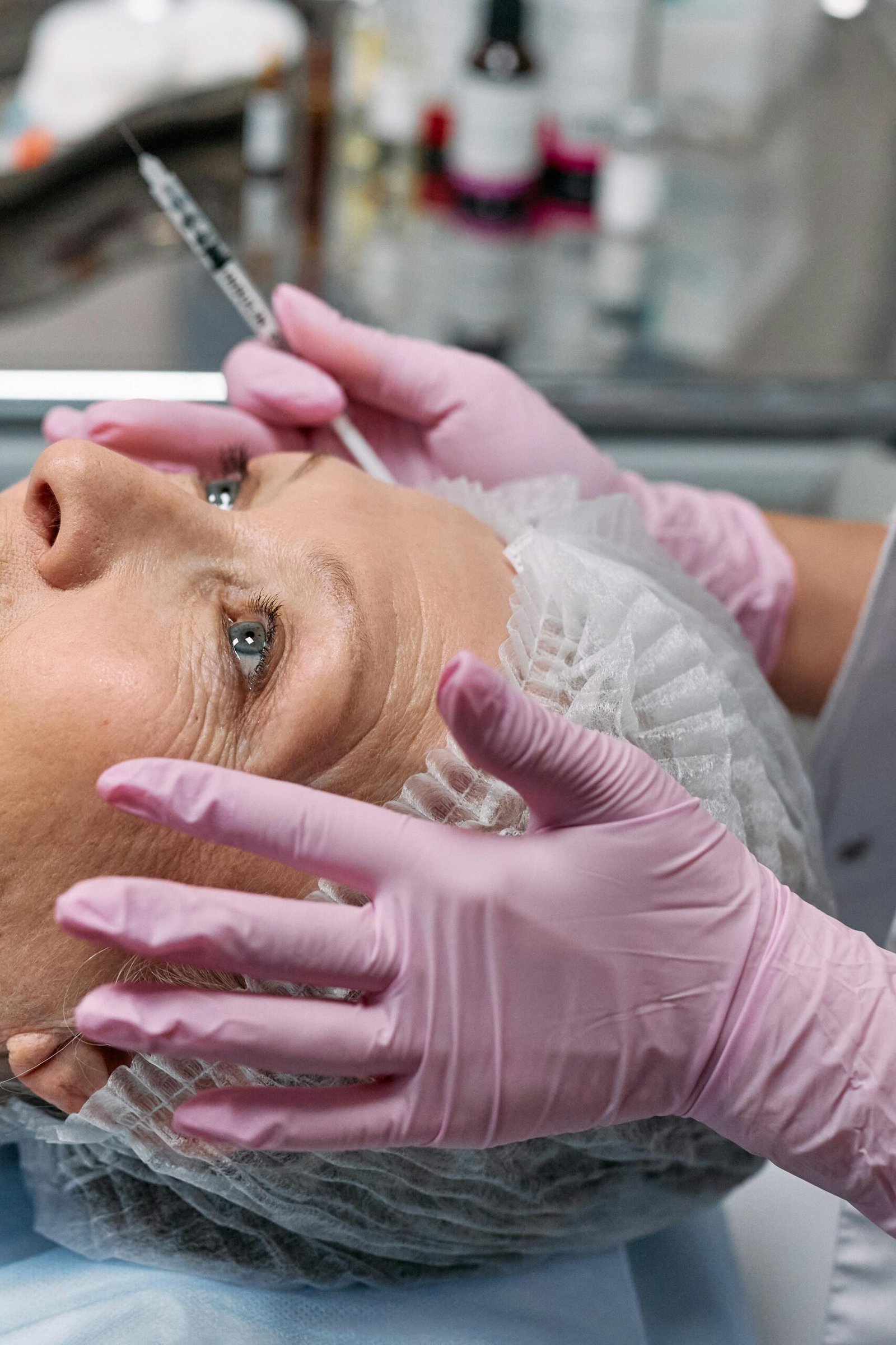 A person receiving a cosmetic injection while lying down, wearing a hairnet. A gloved hand holds a syringe near the forehead.