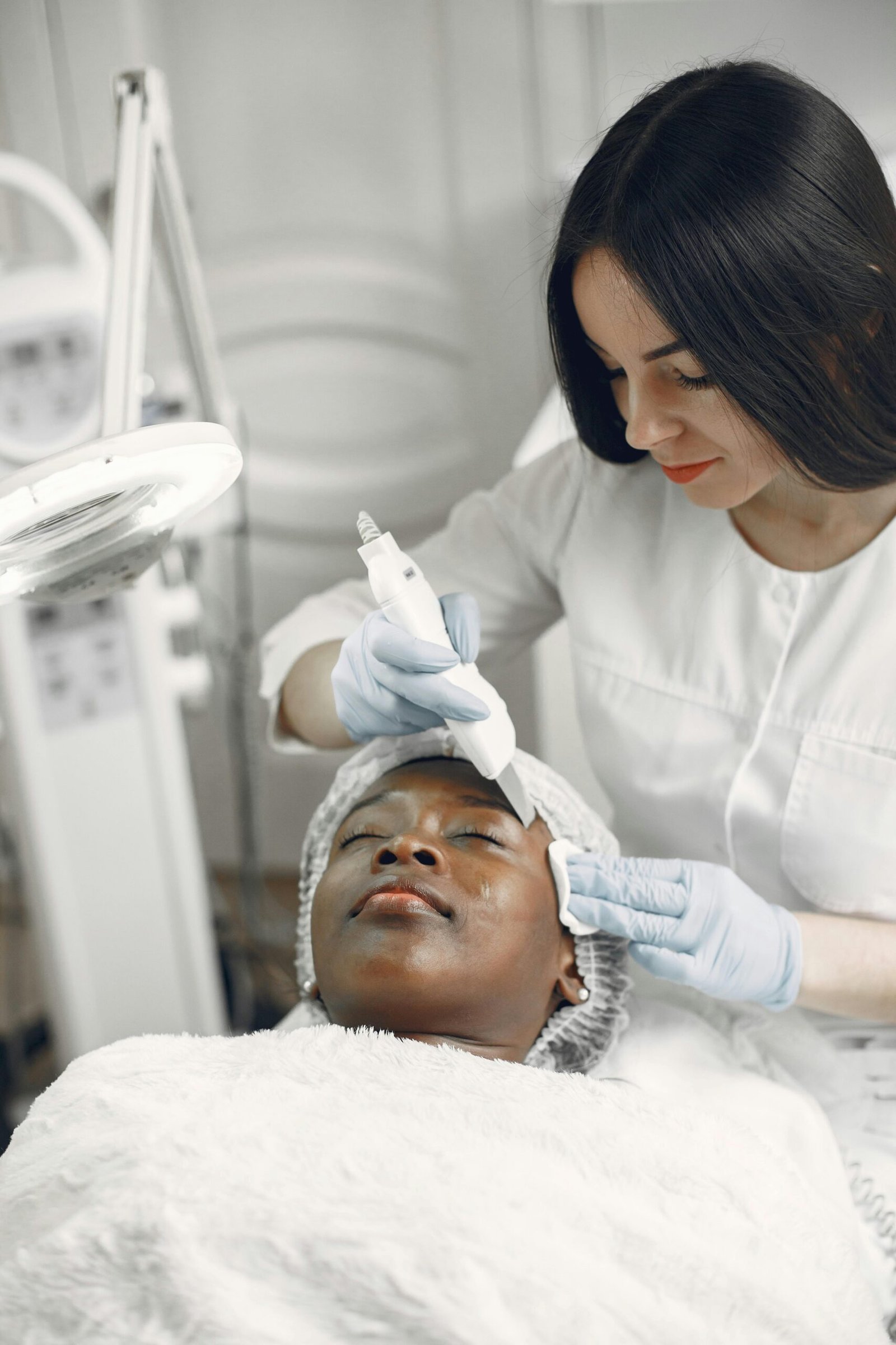 Person receives facial treatment from another in a clinical setting, surrounded by skincare equipment, both wearing white and gloves, creating a professional atmosphere.