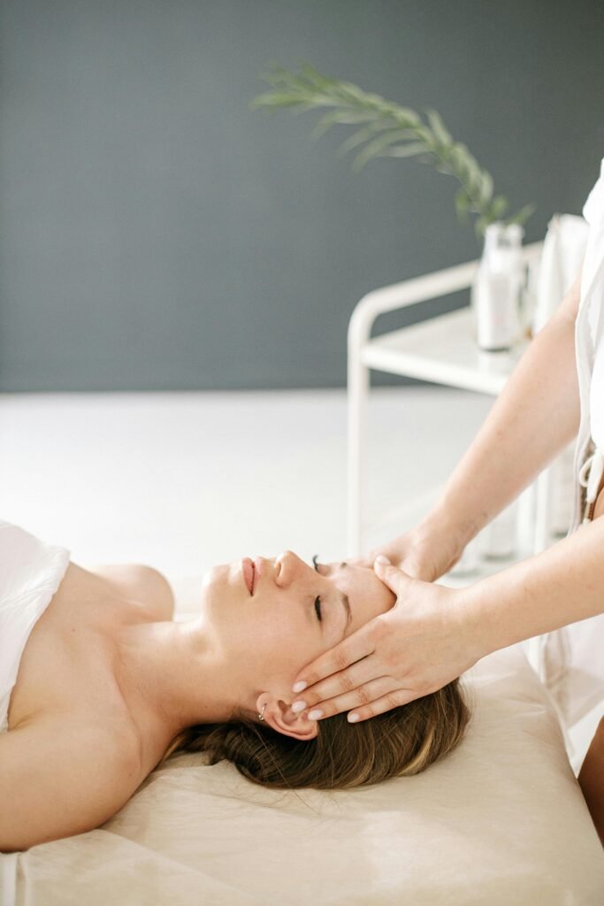 A person receives a relaxing head massage while lying on a bed. Background includes soft lighting and a minimalist decor with plants.