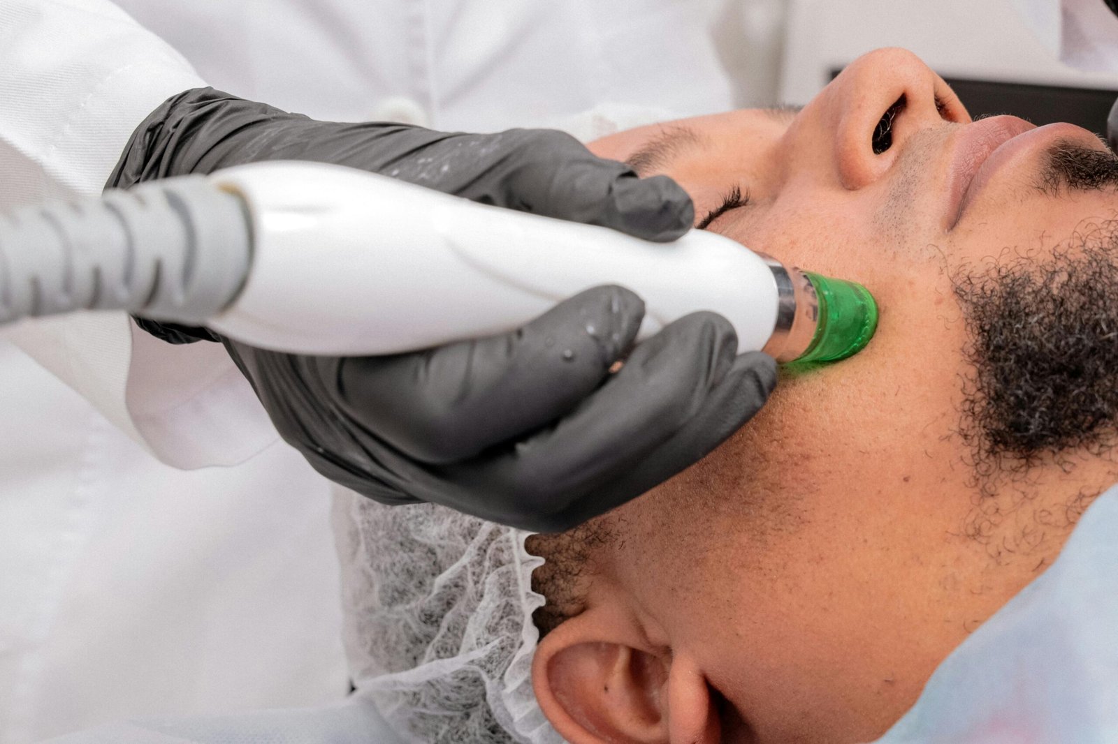 A person receives a facial skin treatment using a device, handled by another person wearing gloves and a white coat.