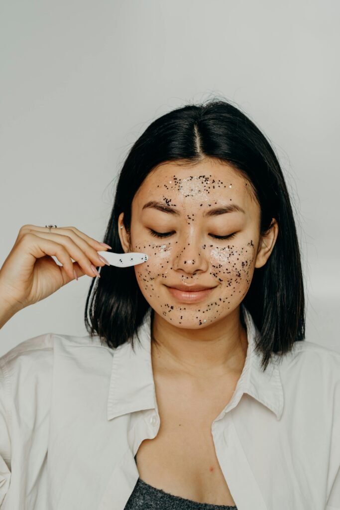 A person with closed eyes applies a facial mask with exfoliating black particles using a small spatula, wearing a white shirt indoors.