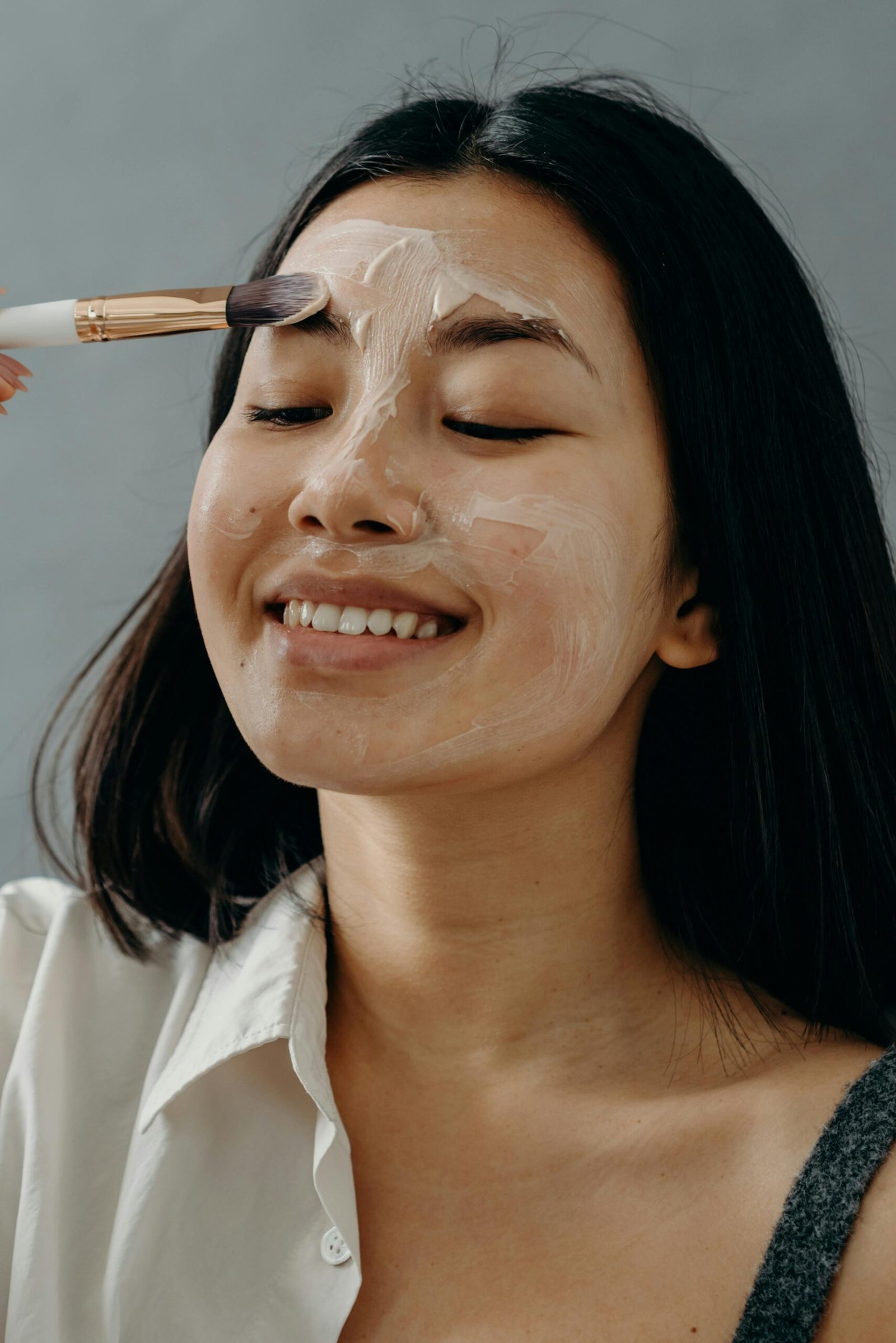 A person is smiling with eyes closed while applying a face mask using a brush against a plain background.