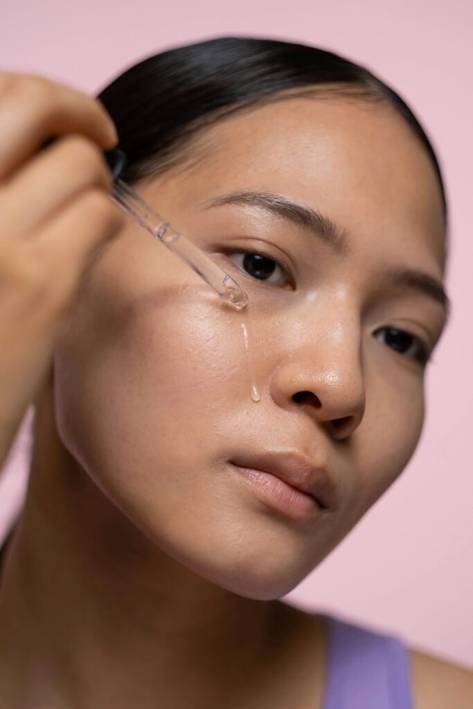 A person applies serum with a dropper to their cheek, against a soft pink background. Close-up skincare routine.