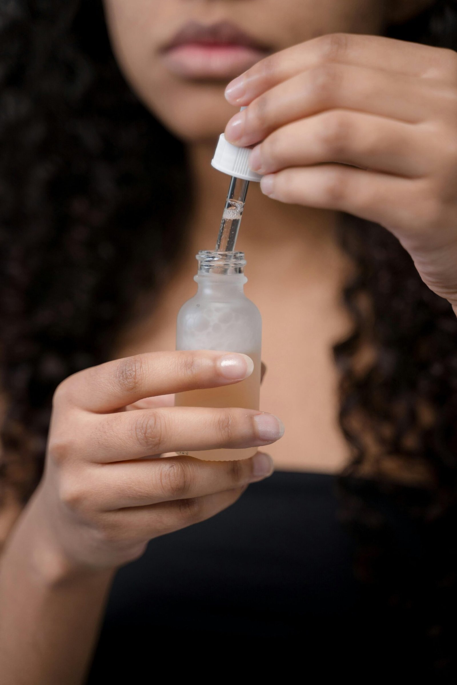 A person with curly hair holds a small dropper bottle, carefully drawing liquid with the dropper against a blurred background.
