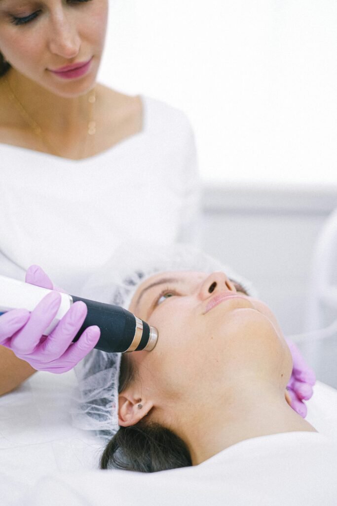 A person wearing gloves performs a skincare treatment on another person's face using a handheld device in a clinical setting.