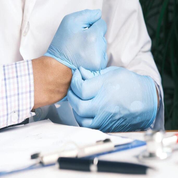A person wearing a lab coat puts on blue surgical gloves. A clipboard and stethoscope are visible on the table.