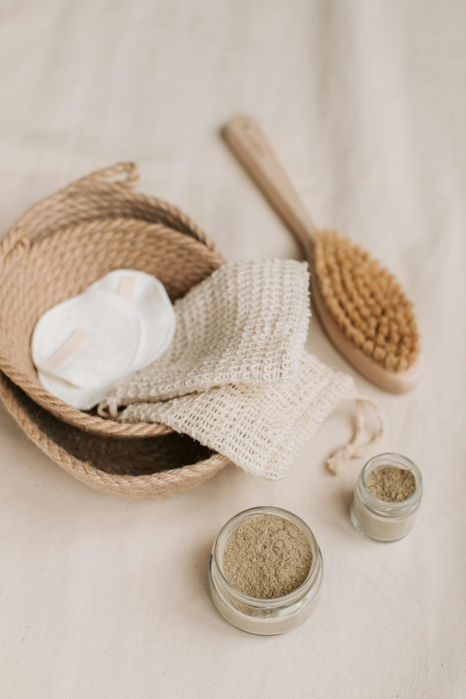 Natural spa setup with woven baskets, wooden brush, reusable pads, cloth, and jars of herbal powder on a beige fabric background.
